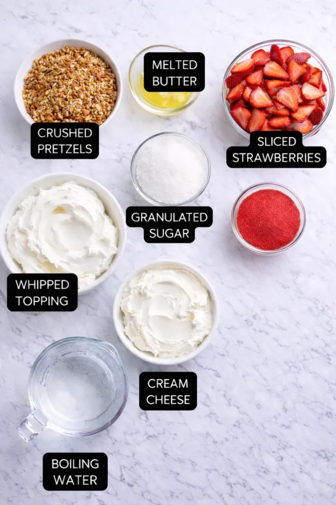Overhead view of strawberry pretzel salad ingredients on a white marble counter with crushed pretzels, melted butter, sliced strawberries, sugar, whipped topping, cream cheese, strawberry gelatin, and boiling water