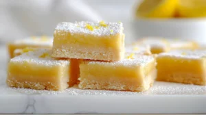 Close-up of homemade lemon bars stacked on a white plate, dusted with powdered sugar and lemon zest, with lemons in the background.