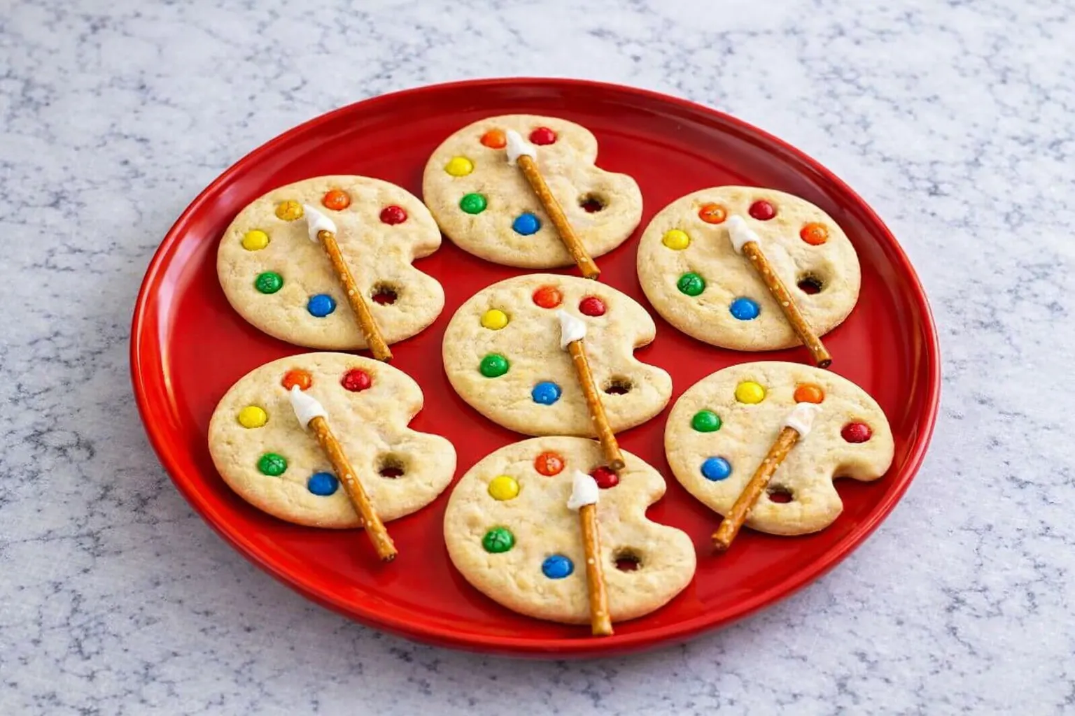 Overhead view of paint palette sugar cookies on a plate, each decorated with colorful M&M’s and a pretzel stick brush tipped with icing.