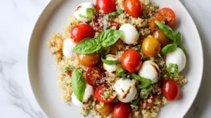 Caprese quinoa salad served on a white plate with cherry tomatoes, mozzarella pearls, basil, and balsamic glaze on a white marble counter