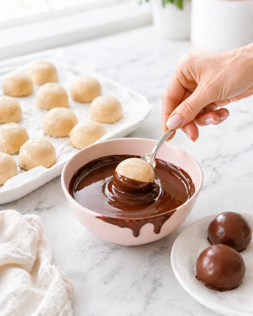 A female hand with a light pink manicure dips a frozen peanut butter yogurt bite into a bowl of melted chocolate on a white marble countertop, with undipped bites on a parchment-lined tray and finished chocolate-coated bites nearby.