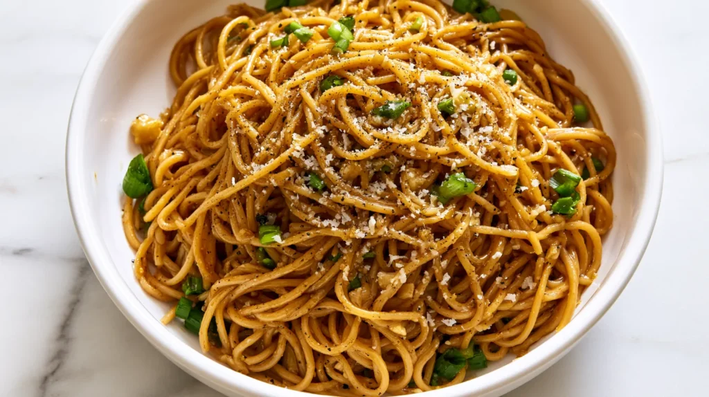 Hyper-realistic overhead food photography of Asian garlic noodles made with long spaghetti-style noodles coated in a glossy warm golden-brown buttery garlic soy-oyster sauce, visible finely minced garlic, sliced green onions, a light sprinkle of finely grated Parmesan, tiny flecks of black pepper, silky sauce clinging to every noodle strand, served in a white shallow ceramic bowl centered on a white marble kitchen counter, noodles arranged in a natural glossy mound, bright white kitchen background, soft natural window light, crisp editorial food photography, realistic texture, clean top-down composition, no hands, no clutter, no text, no watermark