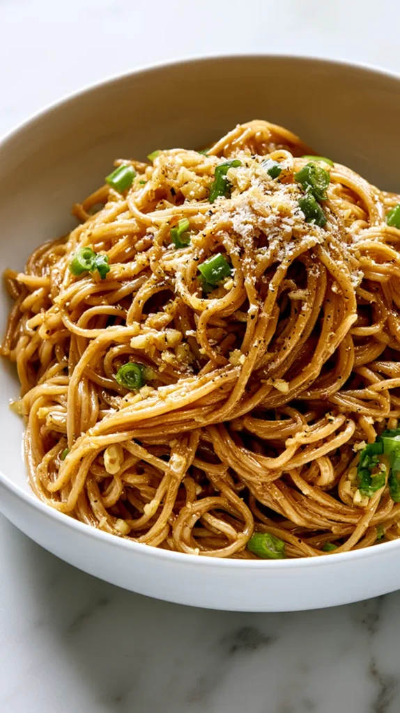 Hyper-realistic overhead food photography of Asian garlic noodles made with long spaghetti-style noodles coated in a glossy warm golden-brown buttery garlic soy-oyster sauce, visible finely minced garlic, sliced green onions, a light sprinkle of finely grated Parmesan, tiny flecks of black pepper, silky sauce clinging to every noodle strand, served in a white shallow ceramic bowl centered on a white marble kitchen counter, noodles arranged in a natural glossy mound, bright white kitchen background, soft natural window light, crisp editorial food photography, realistic texture, clean top-down composition, no hands, no clutter, no text, no watermark