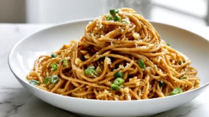 Hyper-realistic hero shot of Asian garlic noodles made with long spaghetti-style noodles coated in a glossy warm golden-brown buttery garlic soy-oyster sauce, visible finely minced garlic, sliced green onions, a light sprinkle of finely grated Parmesan, tiny flecks of black pepper, silky sauce clinging to every noodle strand, served in a white shallow ceramic bowl on a white marble kitchen counter, noodles piled high in a cozy glossy mound, slight steam rising, bright white kitchen background softly blurred, soft natural window light, crisp editorial food photography, realistic texture, clean inviting composition, no clutter, no text, no watermark
