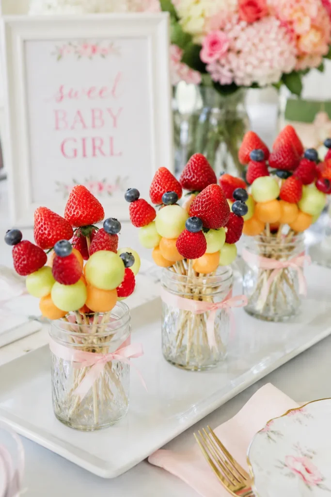 Fruit bouquets with strawberries, blueberries, and melon balls in glass jars with pink ribbons, displayed on white platter beside sweet baby girl sign and pink flower arrangement